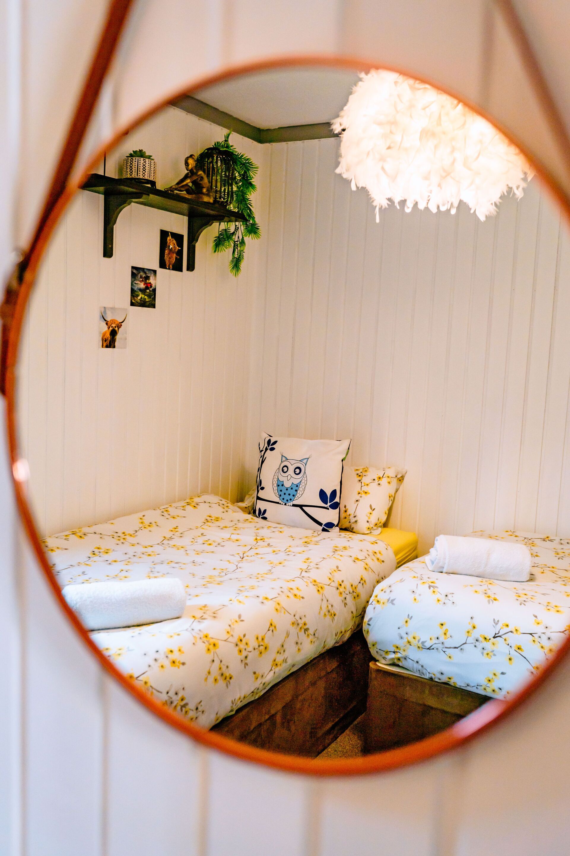 A bedroom with two beds, each with floral bedding, a nightstand, a wall-mounted shelf with plants, and a hanging light fixture.