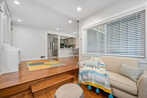 A spacious living area with a wooden floor, a beige sofa, a colorful rug, and a large window with blinds.
