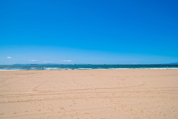 A wide sandy beach with footprints leading to the water, a clear blue sky, and distant ships on the horizon.