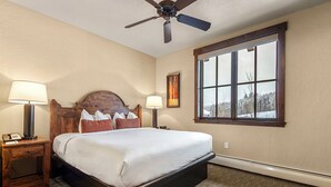 A bedroom with a bed, two bedside tables with lamps, a ceiling fan, and a large window with a view of trees. - Ski-in/out Grand Lodge Peak 7 Suite. (Breckenridge)