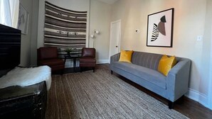 A living room with a grey sofa, two brown chairs, a striped wall hanging, and a framed abstract artwork.