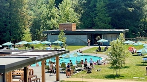 Zona de piscina con sillas de descanso y sombrillas, rodeada de árboles y un edificio de diseño moderno.