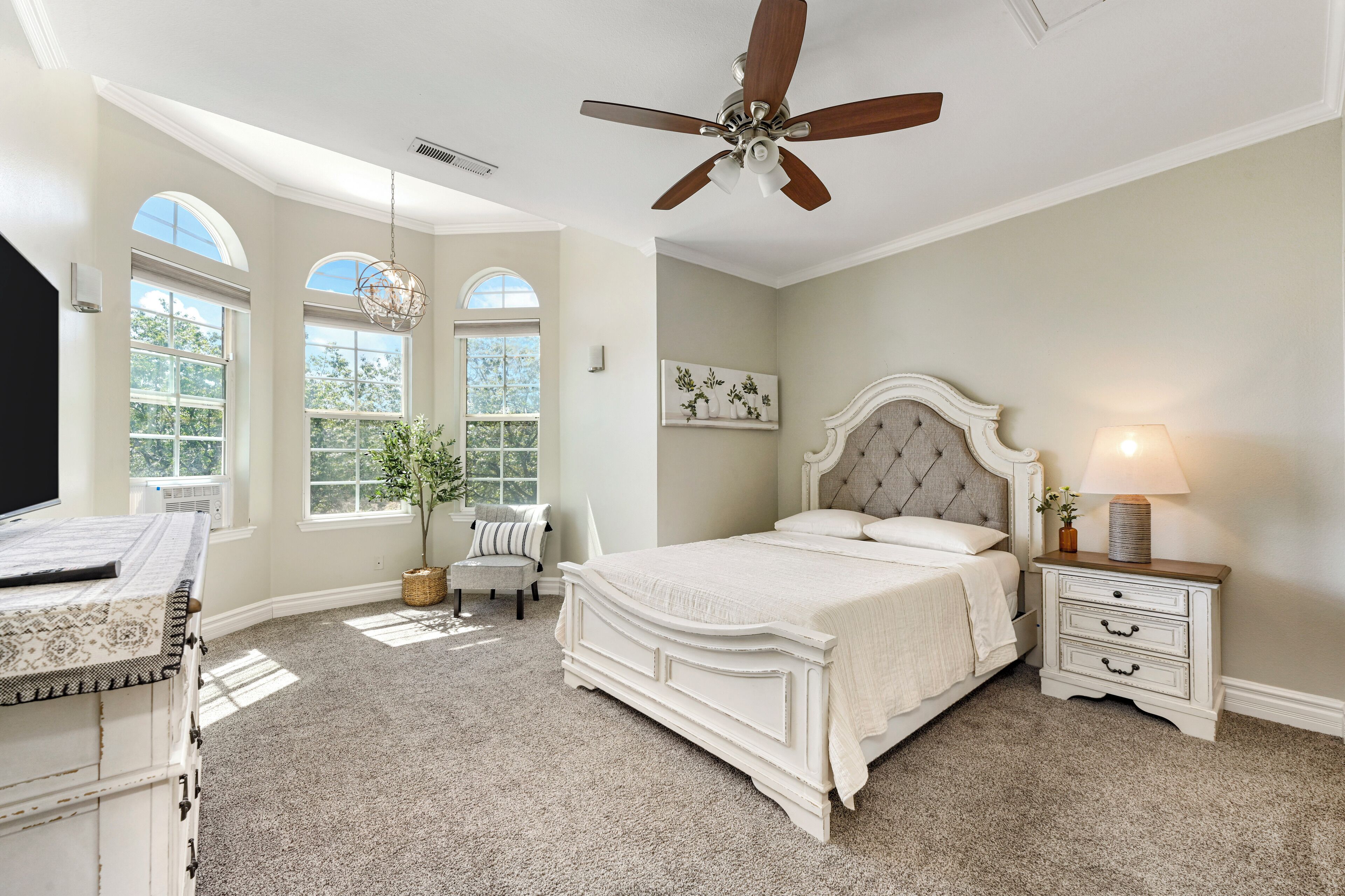 A bedroom with a large bed, a nightstand, a ceiling fan, and a window with a view of trees.