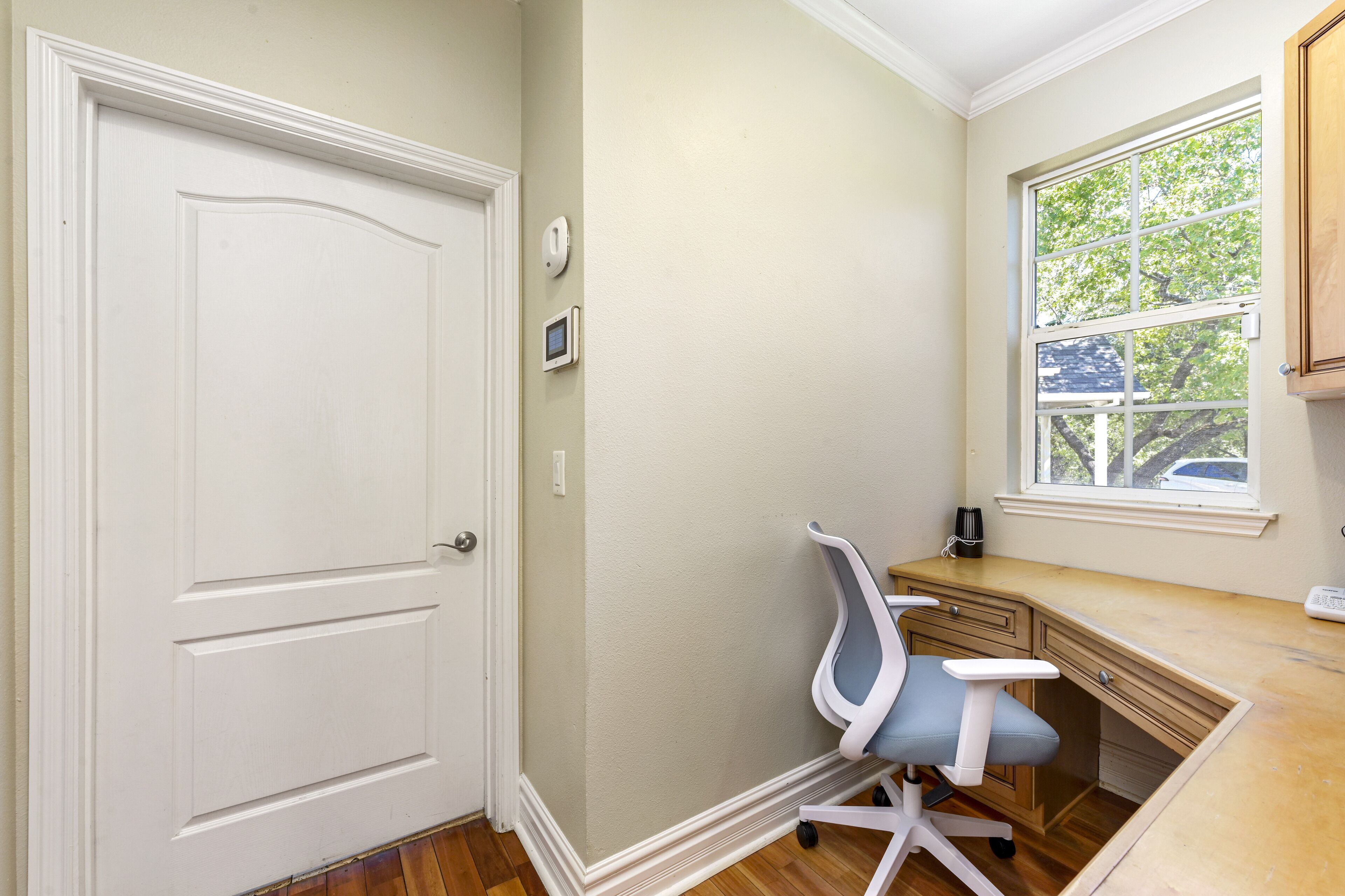 Una habitación con una puerta blanca, un escritorio con una silla, una ventana con vistas a árboles y un termostato en la pared.
