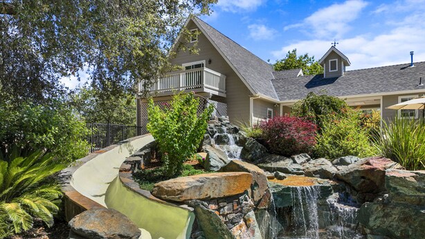 Una propiedad residencial con cascada en el jardín, piscina y una casa con balcón.