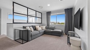 A modern living room with a grey sectional sofa, a small side table, a flat-screen TV mounted on the wall, and a large window with curtains. - Putt Putt - Pool Slide - 100m to Beach - Ultimate Family Entertainer (Kingscliff)