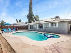 Un patio trasero residencial con piscina, terraza cubierta y un árbol alto.