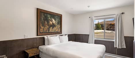 A hotel room with a large bed, a painting of a moose, a window with curtains, and a view of a building outside.