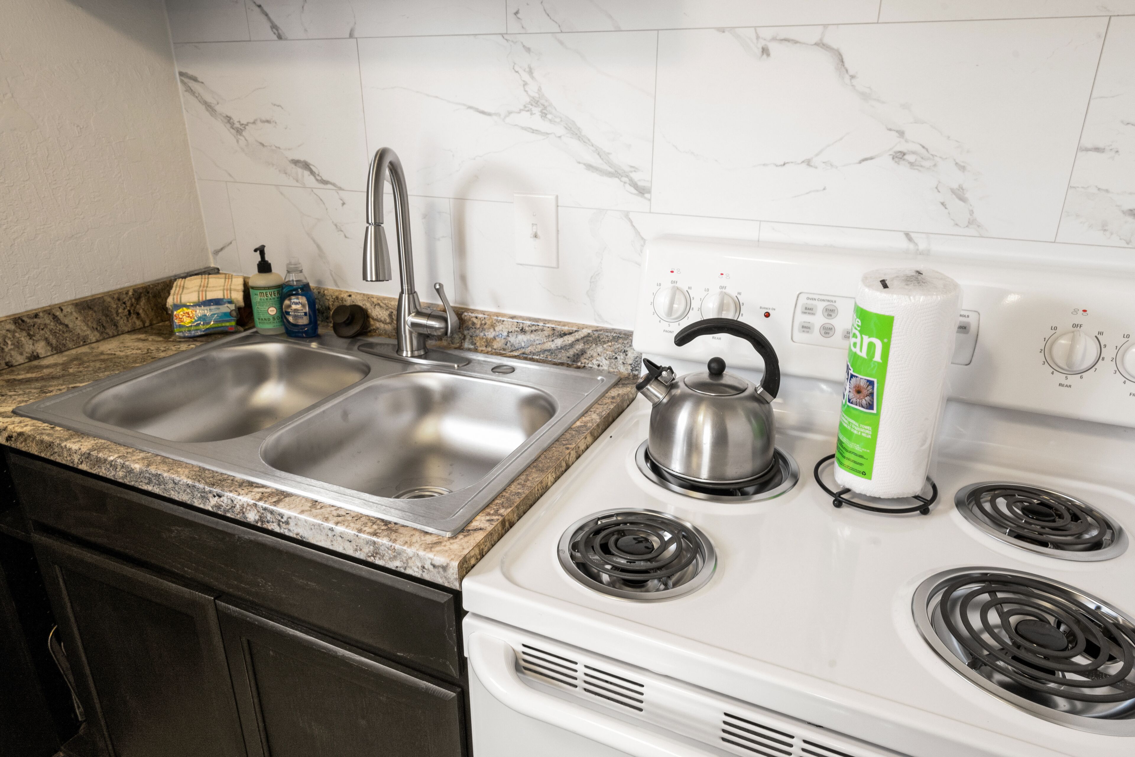 Een keuken met een dubbele spoelbak, een roestvrijstalen kraan, een waterkoker en een fornuis met een keukenrolhouder.