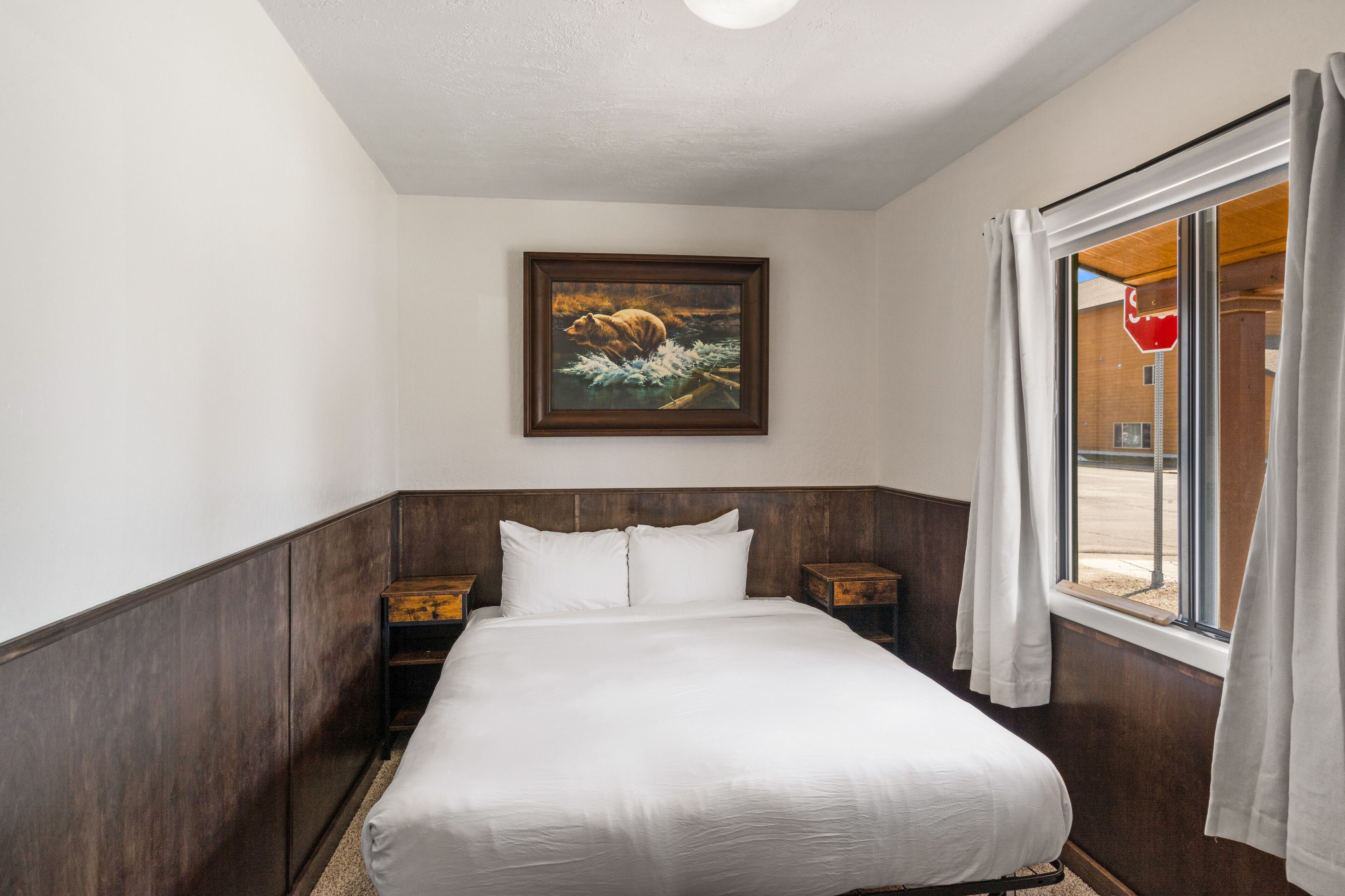 A hotel room with a large bed, wooden headboard, a painting on the wall, and a window with curtains.