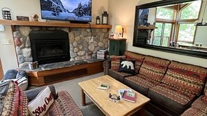 A cozy living room with a stone fireplace, a television mounted above it, and a large window with a view of greenery. - Lola’s Lodge Ski-In/Ski-Out 3 bedroom Lodge near hiking/golf (Lutsen)