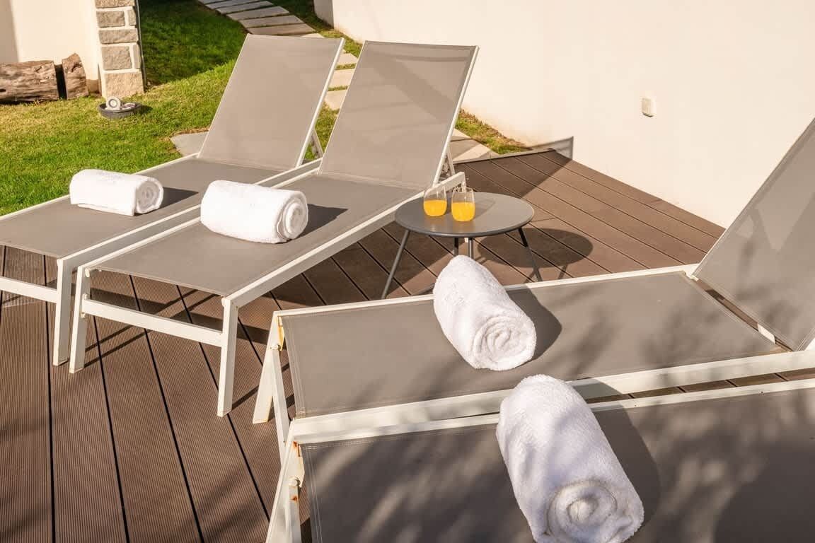 Ein sonniger Außenbereich mit einer Lounge, drei Liegest ühlen, jeweils mit zusammengelegtem Handtuch und einem kleinen Tisch mit einem Glas Orangensaft.