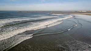Een strand met golven, een pier en een vuurtoren in de verte.