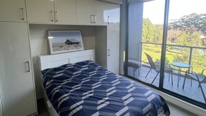 Une chambre à coucher comprenant un lit, une table de chevet, un tableau encadré et une vue sur un balcon aménagé avec du mobilier d’extérieur.