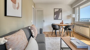 A modern living room with a grey sofa, a dining table with chairs, and a large window offering a city view. - Bright Matthäus 3 Room w/ Storage, near Kleinbasel, by Blueground (Basel)