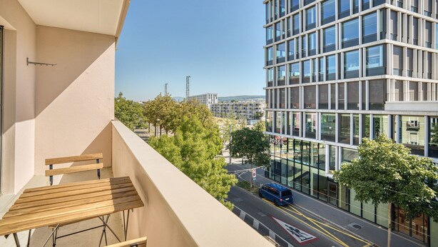 A balcony with a wooden bench overlooking a modern building with large windows and a clear view of the street below. - Bright Matthäus 3 Room w/ Storage, near Kleinbasel, by Blueground (Basel)