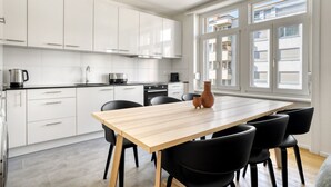 A modern kitchen with white cabinets, a wooden dining table, black chairs, and a large window with a view of buildings. - Upscale Gundelingen 4 Room w/ W/D & Storage, nr Train Station, by Blueground (Basel)