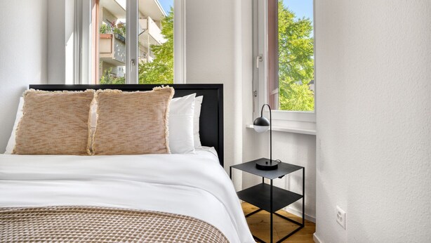 A bedroom with a bed, two pillows, a bedside table, a lamp, and a window with a view of buildings and trees. - Upscale Gundelingen 4 Room w/ W/D & Storage, nr Train Station, by Blueground (Basel)
