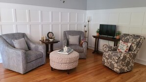 Een woonkamer met drie fauteuils met een patroon, een rond voetenbankje, een tafeltje met een klok en kaarsen, en een televisie op een dressoir.
