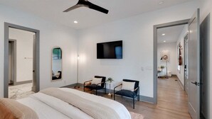 Une chambre moderne avec un grand lit, un ventilateur de plafond, un téléviseur fixé au mur et une petite table avec des chaises.