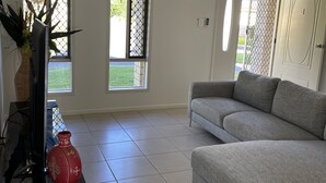 Een woonkamer met een grijze bank, een televisie op een zwart meubelstuk, en een rode vaas. Er zijn twee ramen met metalen tralies en een deur met een codeerapparaat.