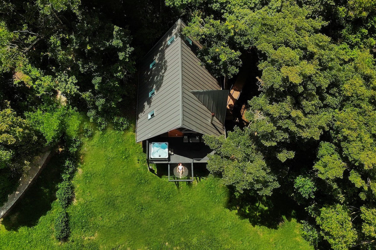 An aerial view of a house surrounded by dense trees, featuring a large swimming pool and a hot tub.
