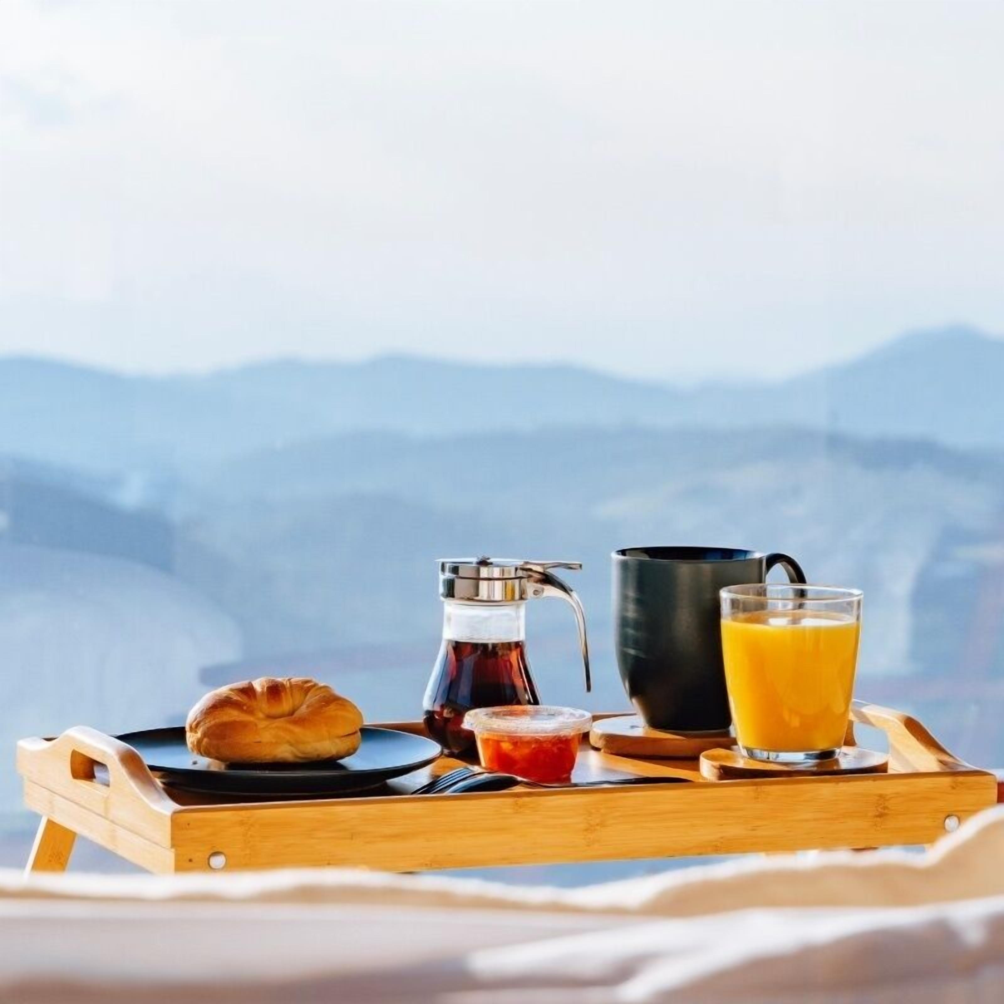 A breakfast tray with a croissant, a coffee pot, a mug, and a glass of orange juice, set against a mountainous backdrop.
