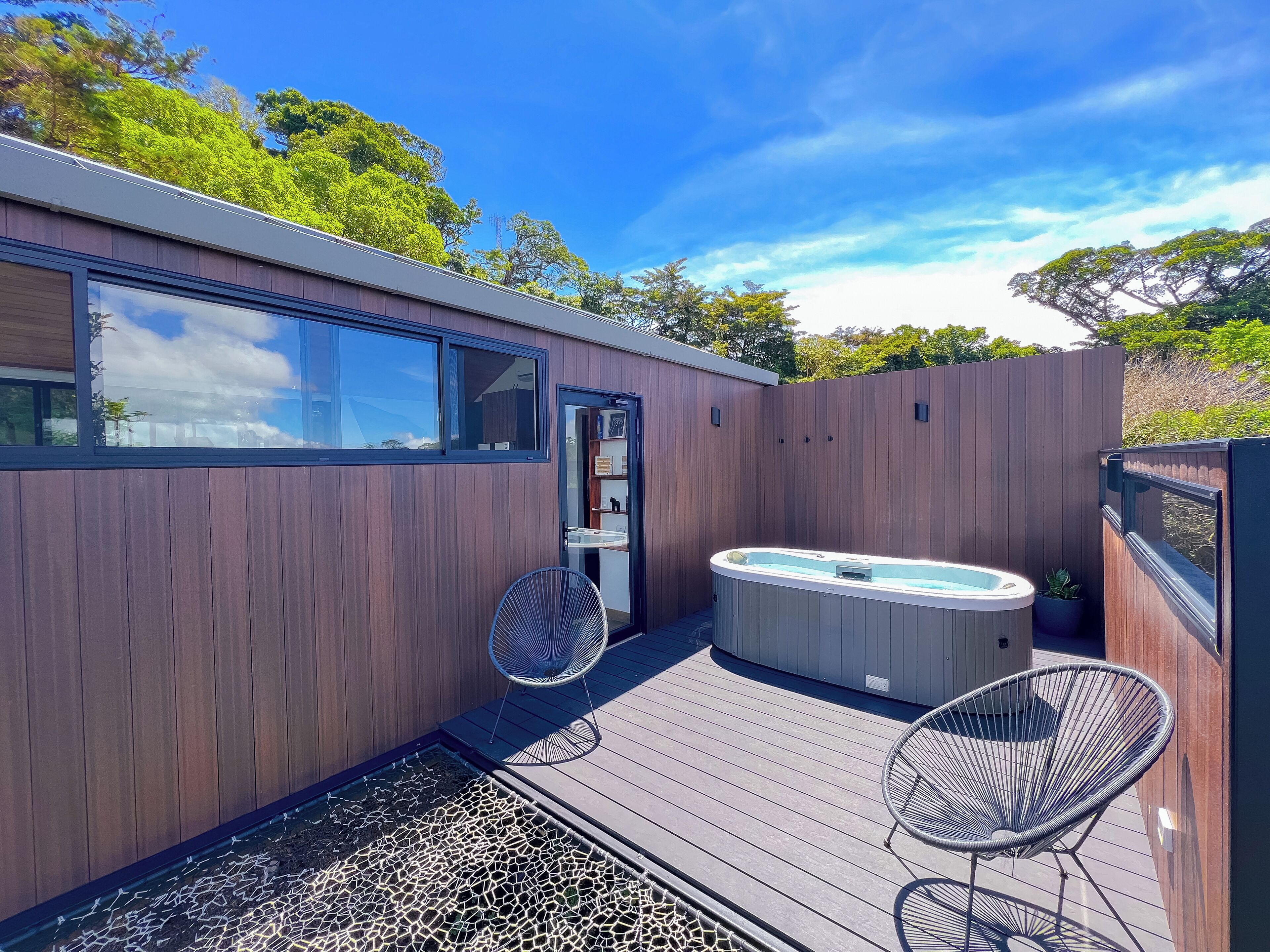 A modern outdoor hot tub area with wooden decking, two wire-framed chairs, and a glass door leading to a room.