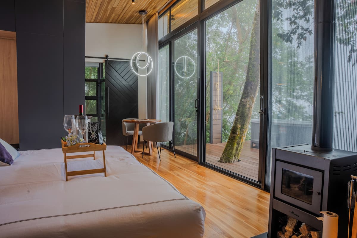 A modern bedroom with a large bed, a wooden side table with glasses and flowers, a small table with chairs, and a wood-burning stove.