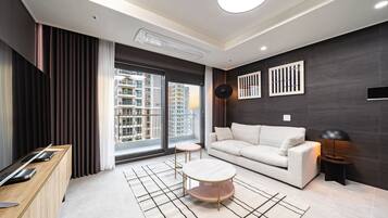 A modern living room with a sofa, a coffee table, a desk, and a large window with a view of high-rise buildings.