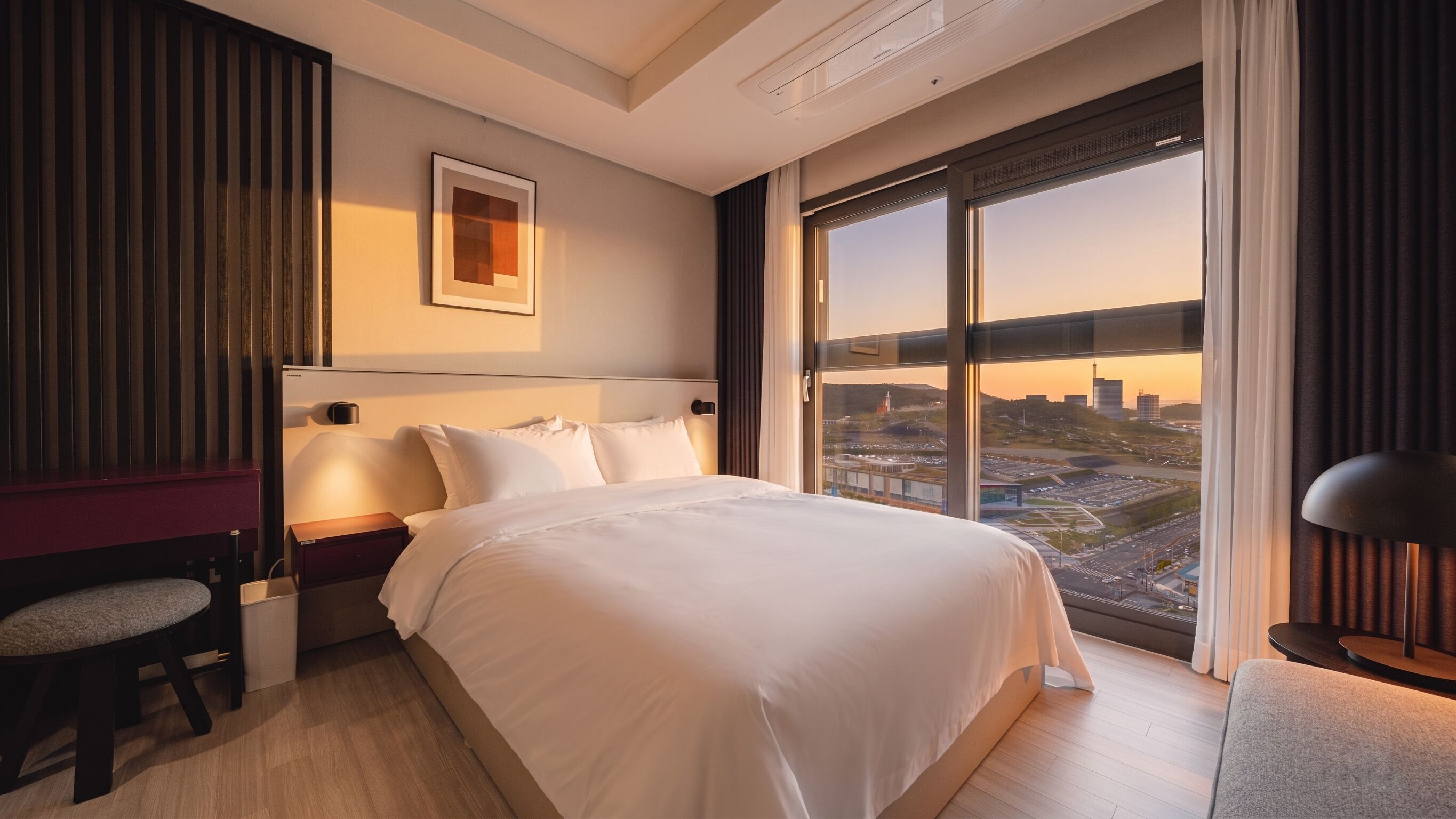 Quarto de hotel com uma cama grande, uma escrivaninha, uma cadeira, uma mesa de cabeceira e vista para a paisagem urbana através de uma grande janela.