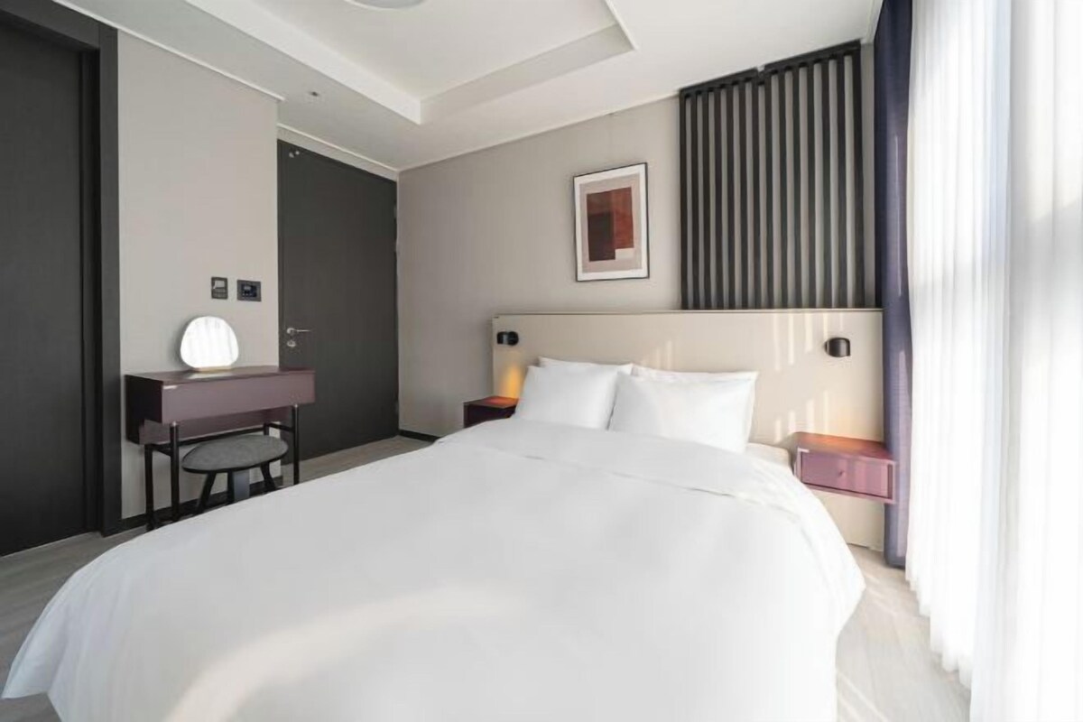 A modern hotel room with a large bed, a desk with a chair, a bedside table with a lamp, and a framed picture on the wall.