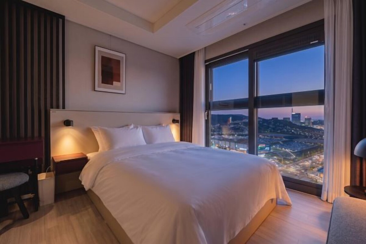 A hotel room with a large bed, a nightstand, a chair, a framed picture on the wall, and a city view through large windows.