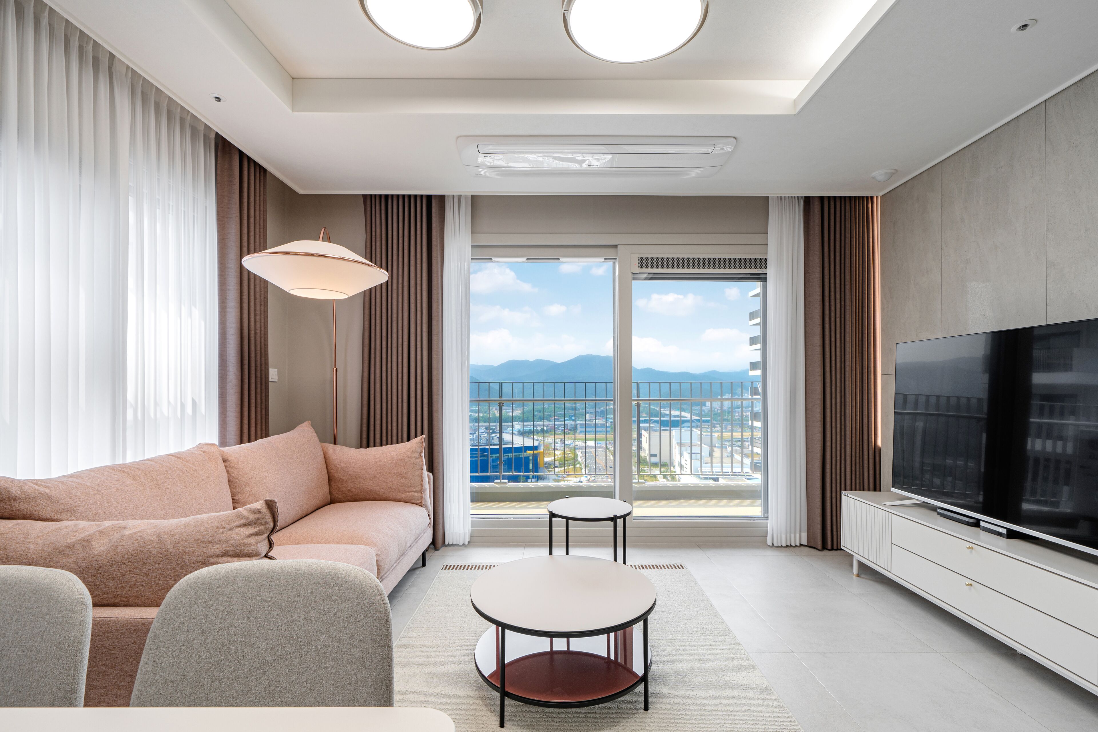 Ett modernt hotellrum med en soffa, en tv, ett litet bord och utsikt över staden genom stora fönster.