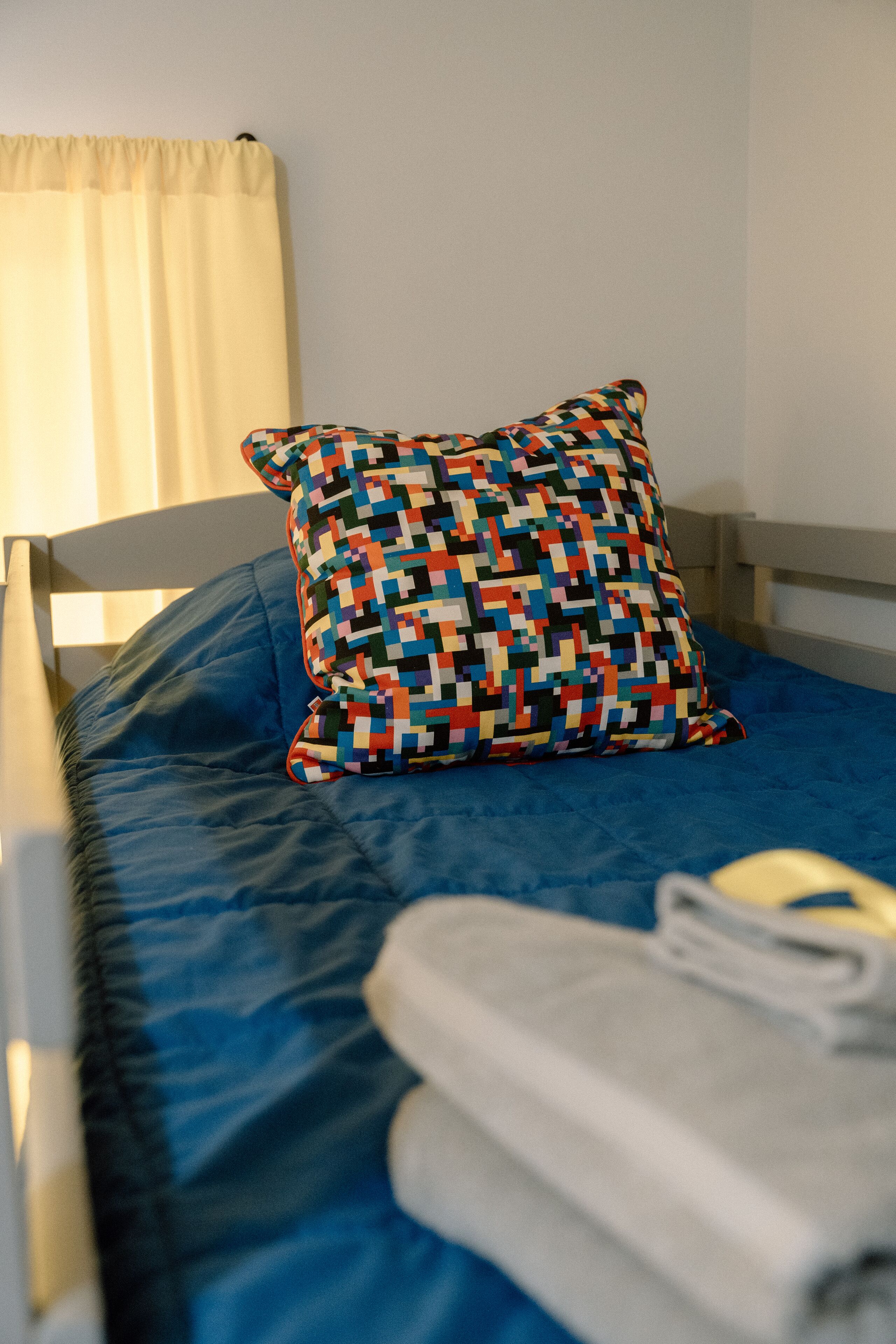 A bed with a blue mattress, a multicolored pillow, and a rolled-up towel.