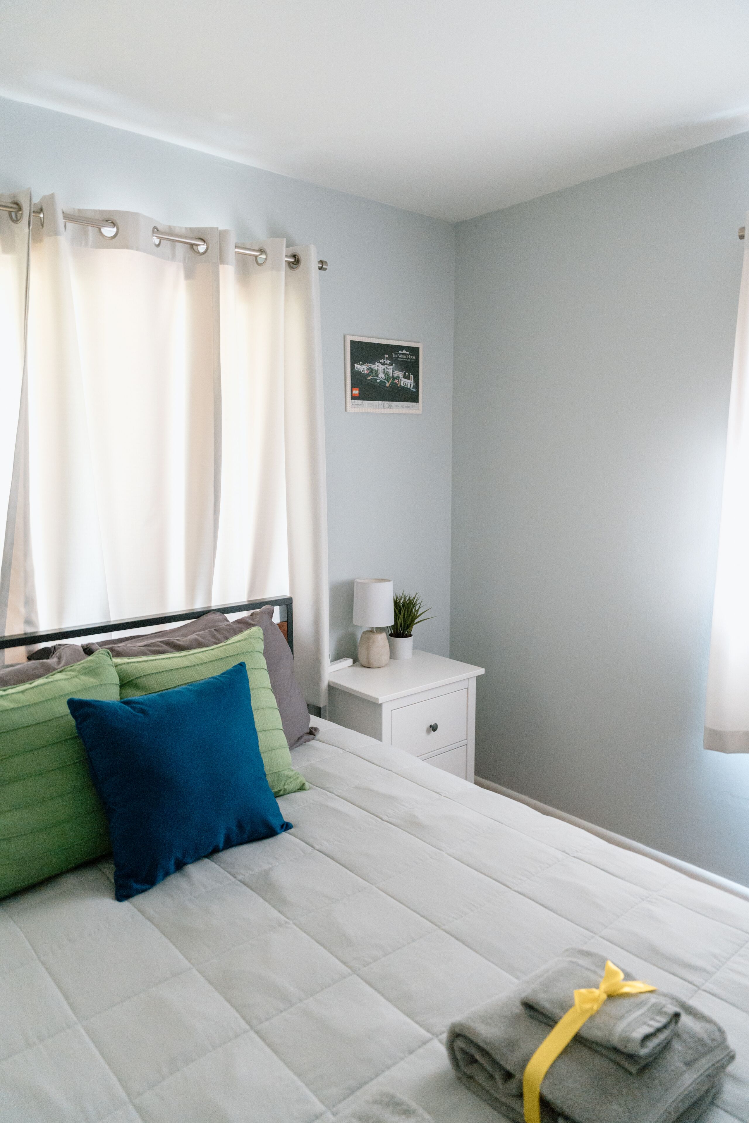 A neatly made bed with pillows, a bedside table with a lamp and a plant, and a framed picture on the wall.