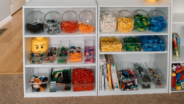 A room with a white wall displaying six framed drawings, a white shelf with various LEGO pieces, and a white cabinet with toys. - Builditplace! Kid friendly Stay, Play and Create! (Akron)