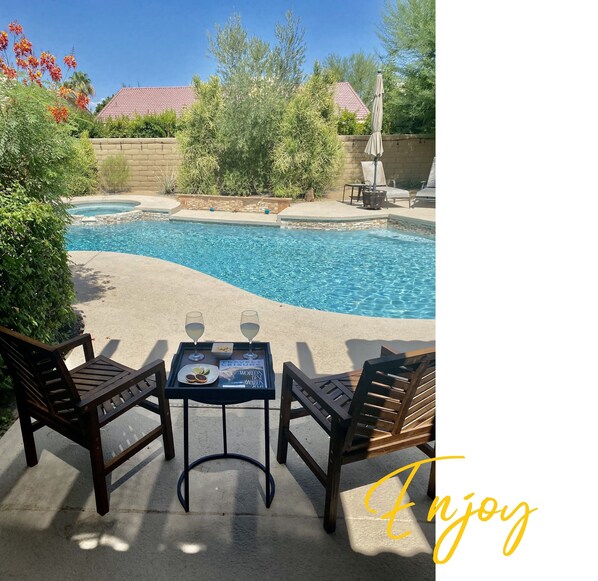 Relax by the serene pool surrounded by lush greenery and a clear sky, with comfortable lounge chairs and a nearby table for a refreshing break.