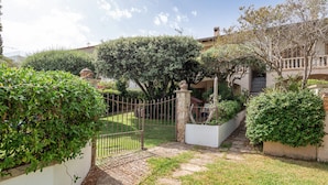 Un jardin clos, accessible par un sentier en pierre menant à l’entrée d’un bâtiment, et entouré d’une végétation luxuriante et d’un mur blanc.