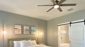 Une chambre à coucher comprenant un lit, un ventilateur de plafond, des lampes fixées au mur et une porte donnant accès à une salle de bain.