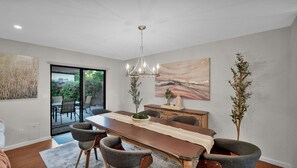 Een eetruimte met een lange houten tafel, grijs gestoffeerde stoelen, een kroonluchter en een groot abstract schilderij aan de muur.