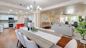 Une cuisine moderne dotée d’appareils électroménagers intégrés, un coin repas avec une table en bois et des chaises blanches, et un salon avec un canapé et un ventilateur de plafond.