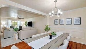 Un espace repas avec une longue table, des chaises blanches, un lustre et des tableaux encadrés accrochés au mur.