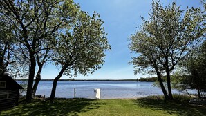 A grassy area with a wooden dock extending into a calm lake, surrounded by trees and a small cabin. - Lakefront Cottage on South Manistique Lake (curtis)