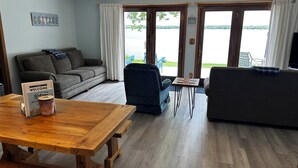 A cozy living room with a wooden table, a sofa, and a blue armchair. There are two doors leading to a balcony with a view of a lake. - Lakefront Cottage on South Manistique Lake (curtis)