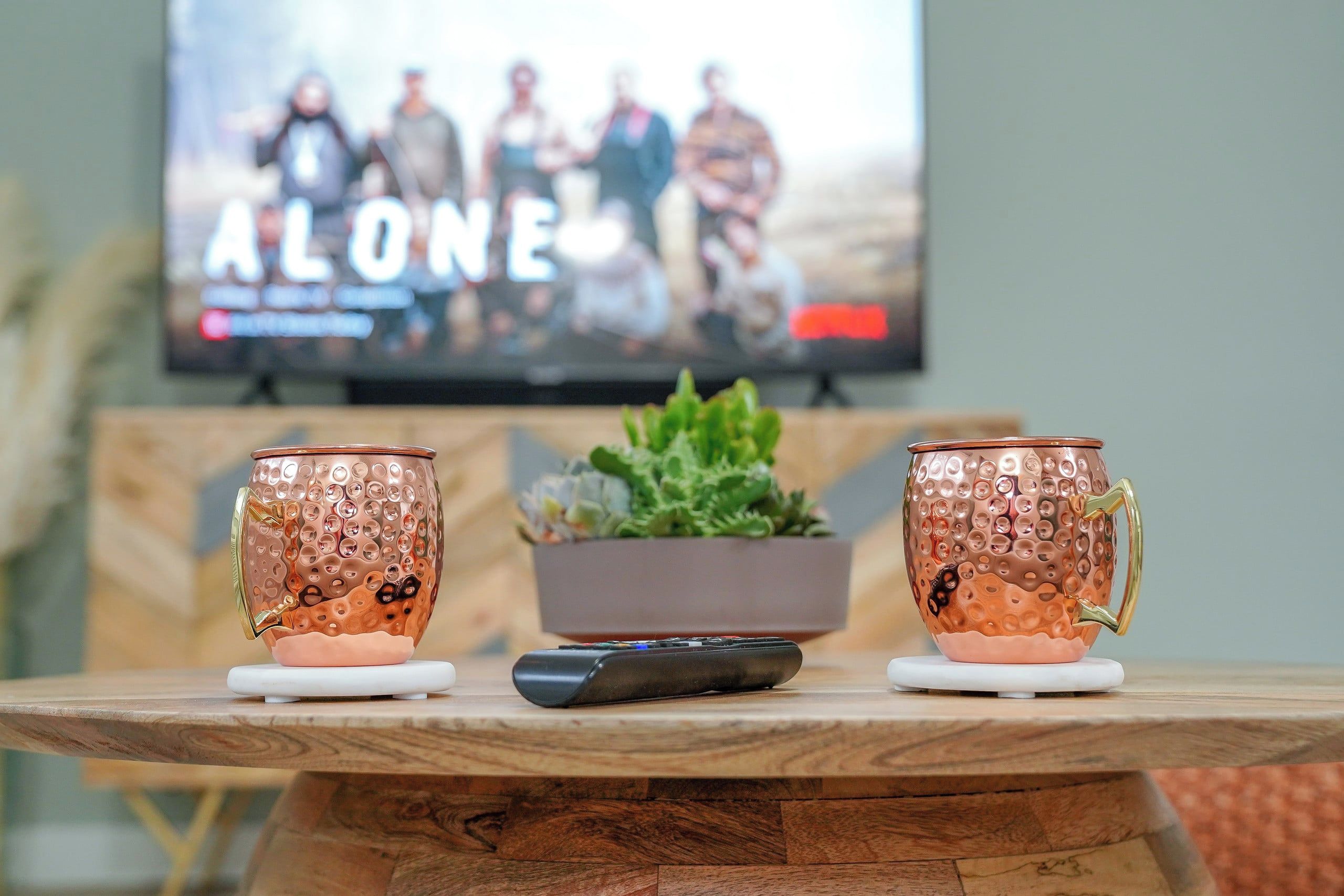 A wooden table with two copper mugs and a remote control. In the background, a television displays a movie scene with the word 'ALONE' visible.