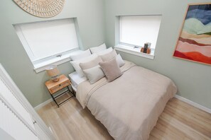 Un dormitorio con una cama, dos ventanas, una mesita de noche y una obra de arte en la pared.