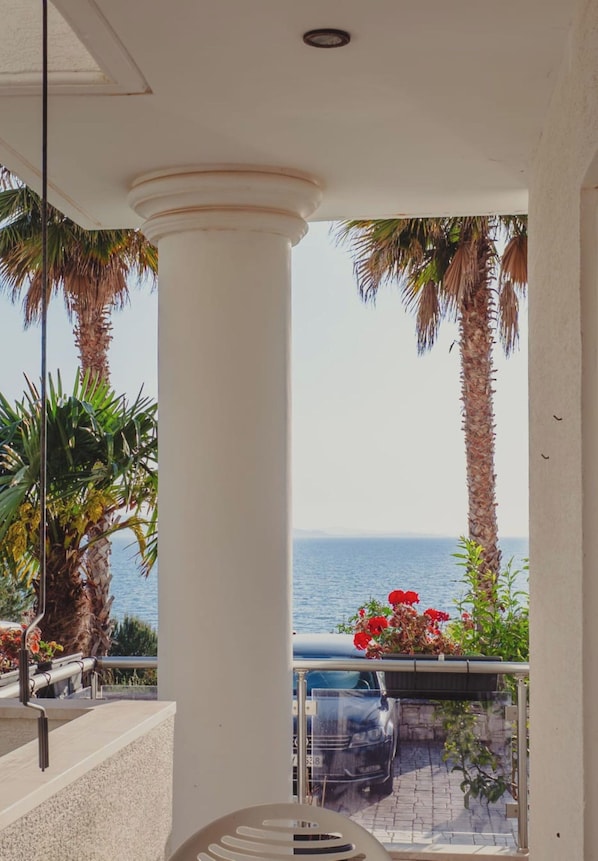 A balcony with a view of the sea, palm trees, and a car parked outside.