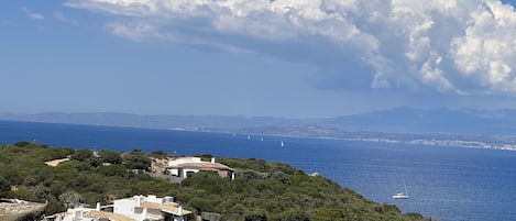 A coastal landscape with a white building surrounded by greenery, overlooking the sea with sailboats and distant mountains.