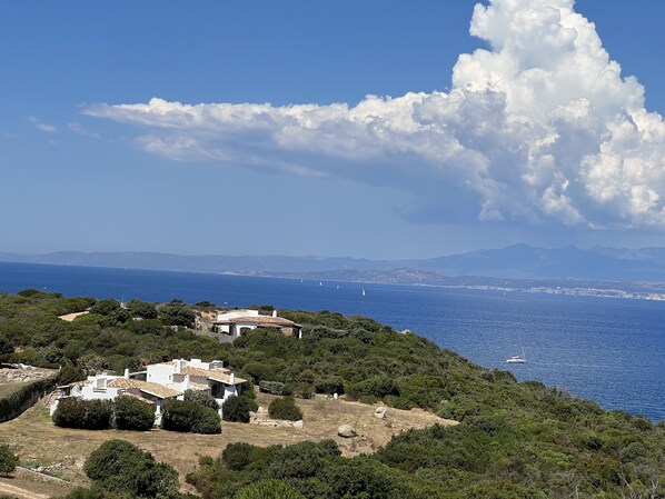 A coastal landscape with a white building surrounded by greenery, overlooking the sea with sailboats and distant mountains.